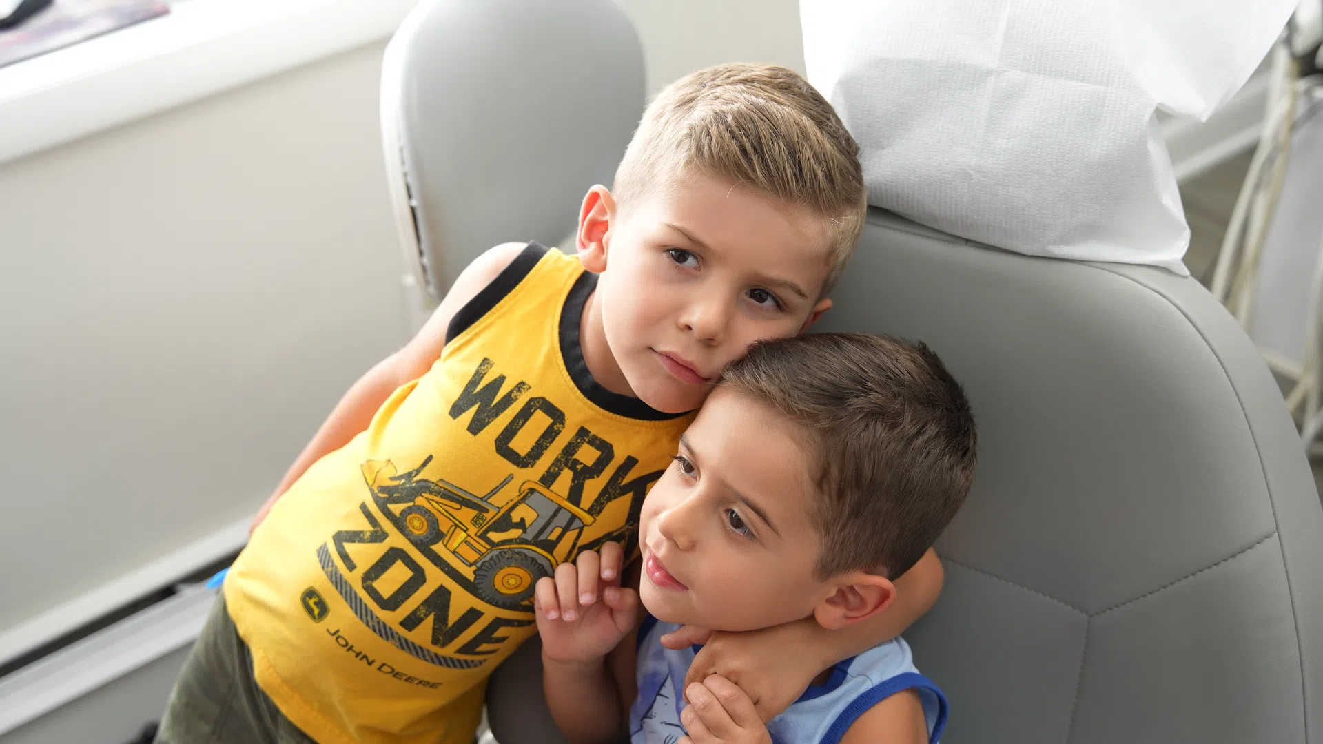 Two young boys sitting closely together showing affection in a bright room near a gray chair.