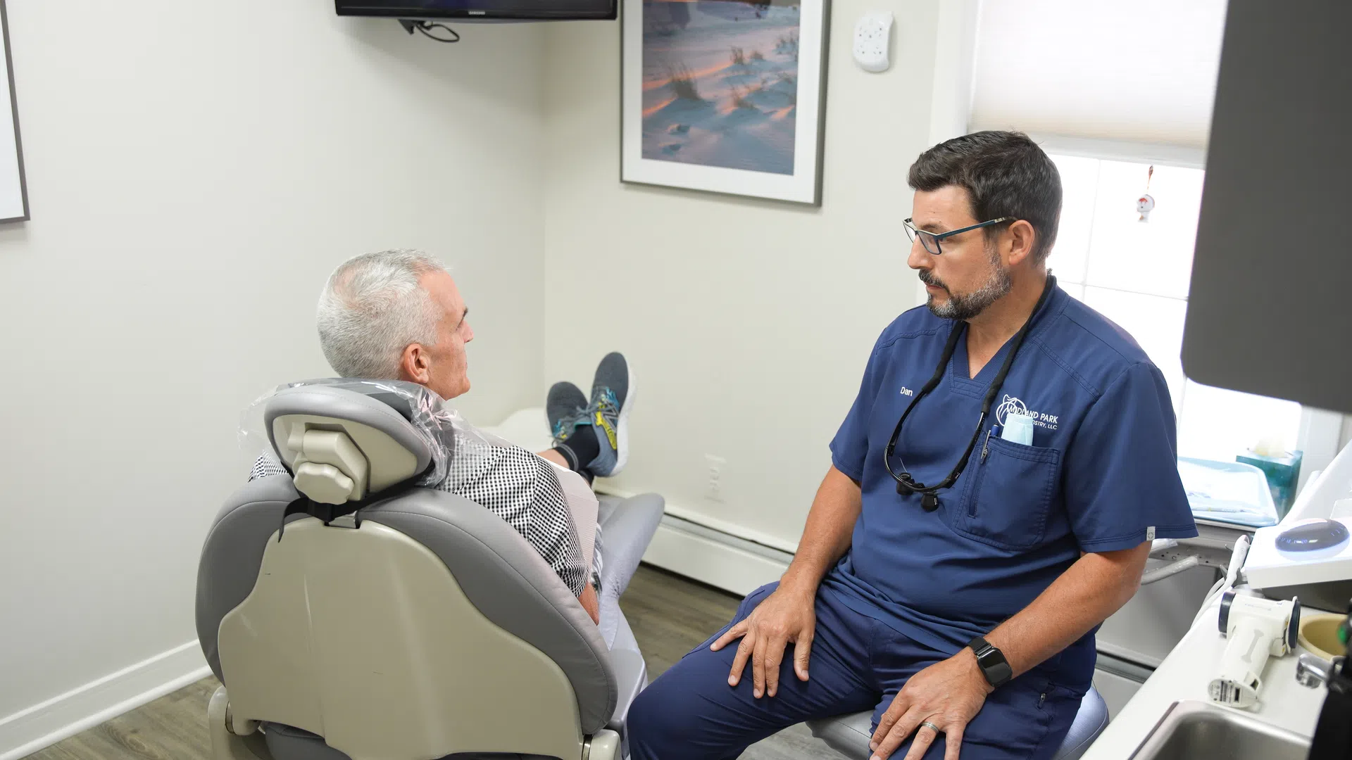 Dentist in blue scrubs consulting with an older male patient seated in a dental chair in a bright clinic room.