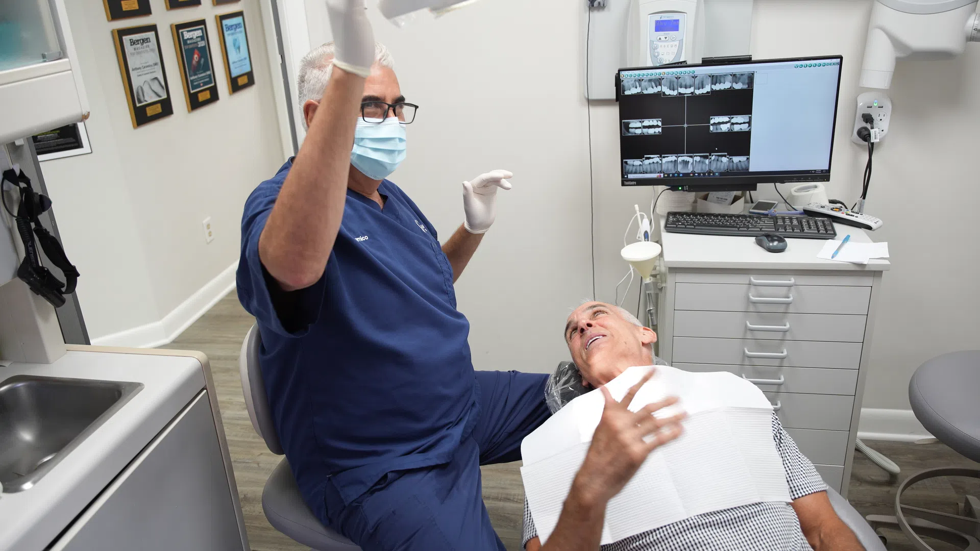 Dentist wearing mask and gloves discusses treatment with patient in dental office with x-ray images.