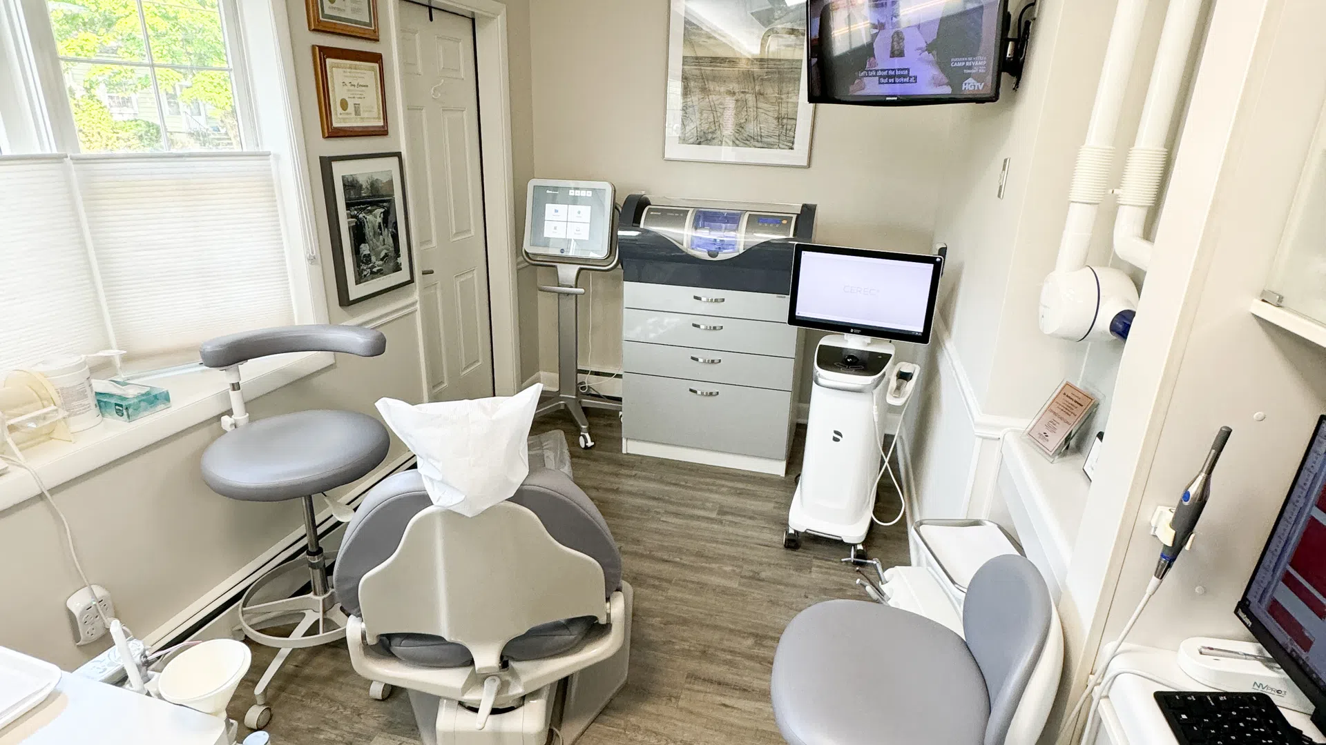 Modern dental clinic room with chair, monitor screens, medical equipment, and natural light from a window.
