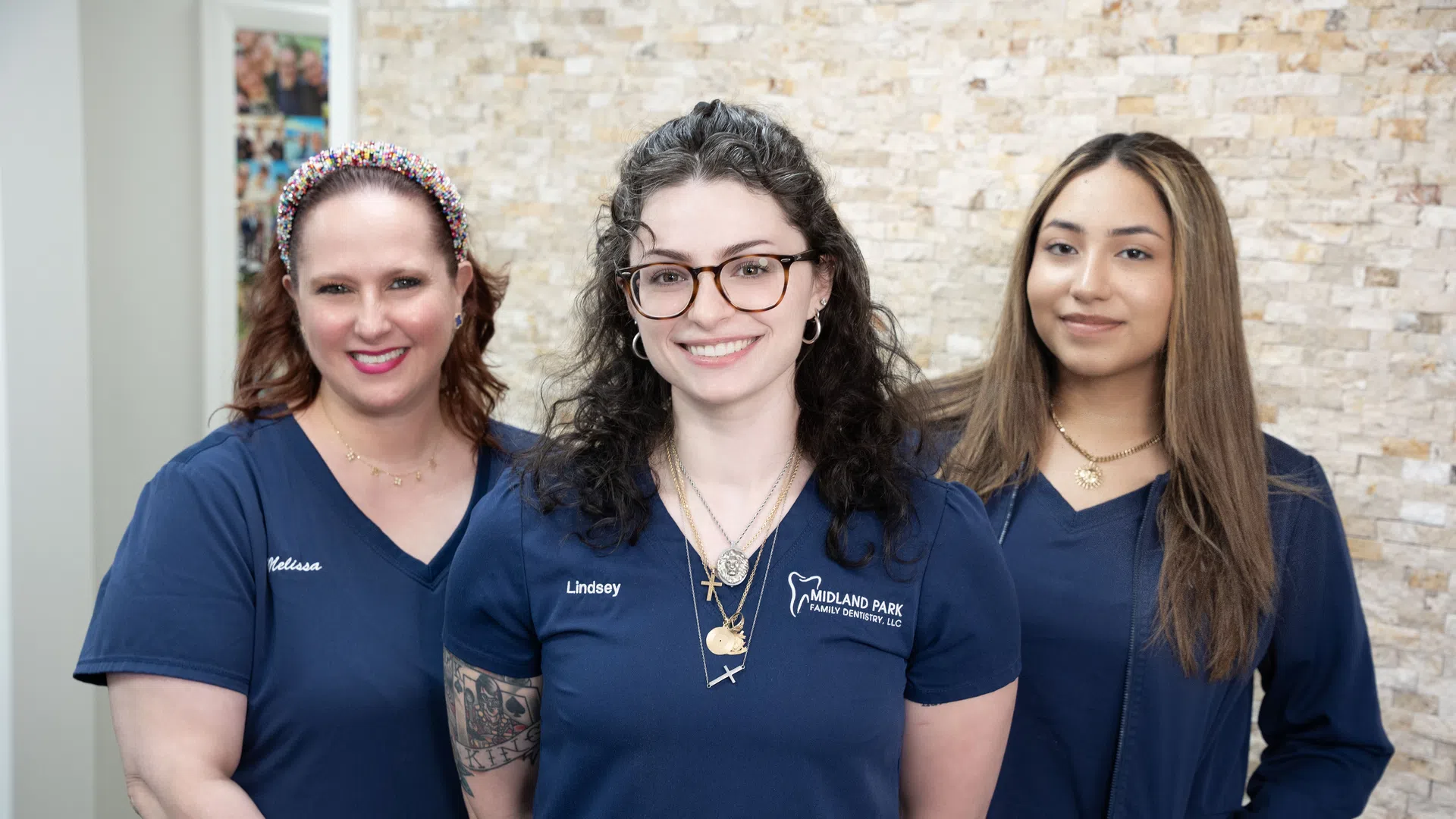Three female dental professionals in blue uniforms smiling indoors against a stone wall background