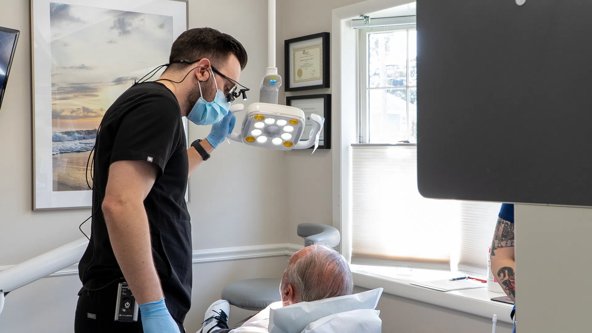 Dentist wearing mask and gloves adjusting dental light over elderly patient in bright dental office.