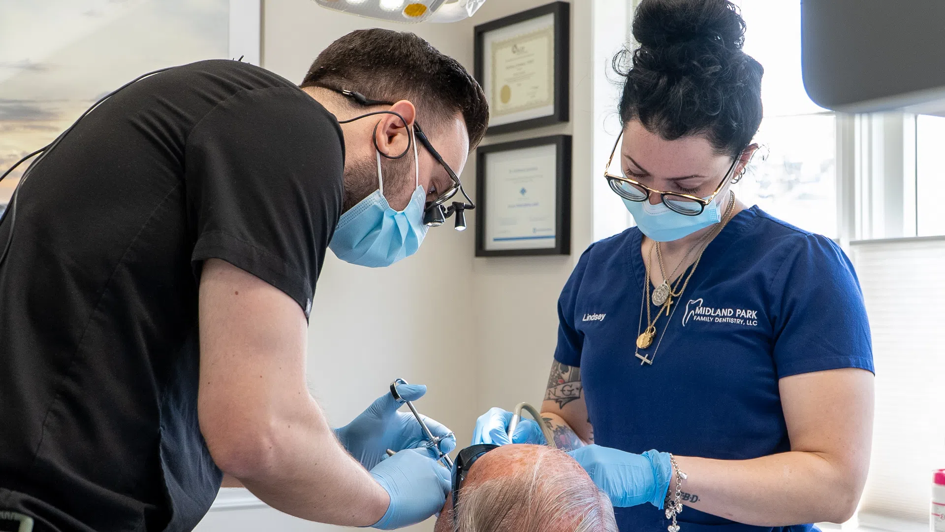 Dentist and dental assistant wearing masks and gloves performing a procedure on a patient in a bright clinic.