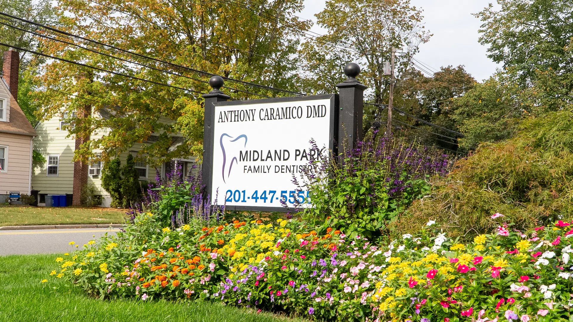 Midland Park Family Dentistry sign surrounded by colorful flowers and greenery near a residential street.