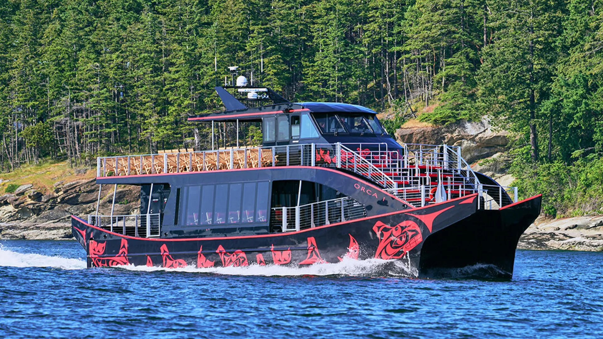 A decorated black boat glides through blue water surrounded by lush green forests.