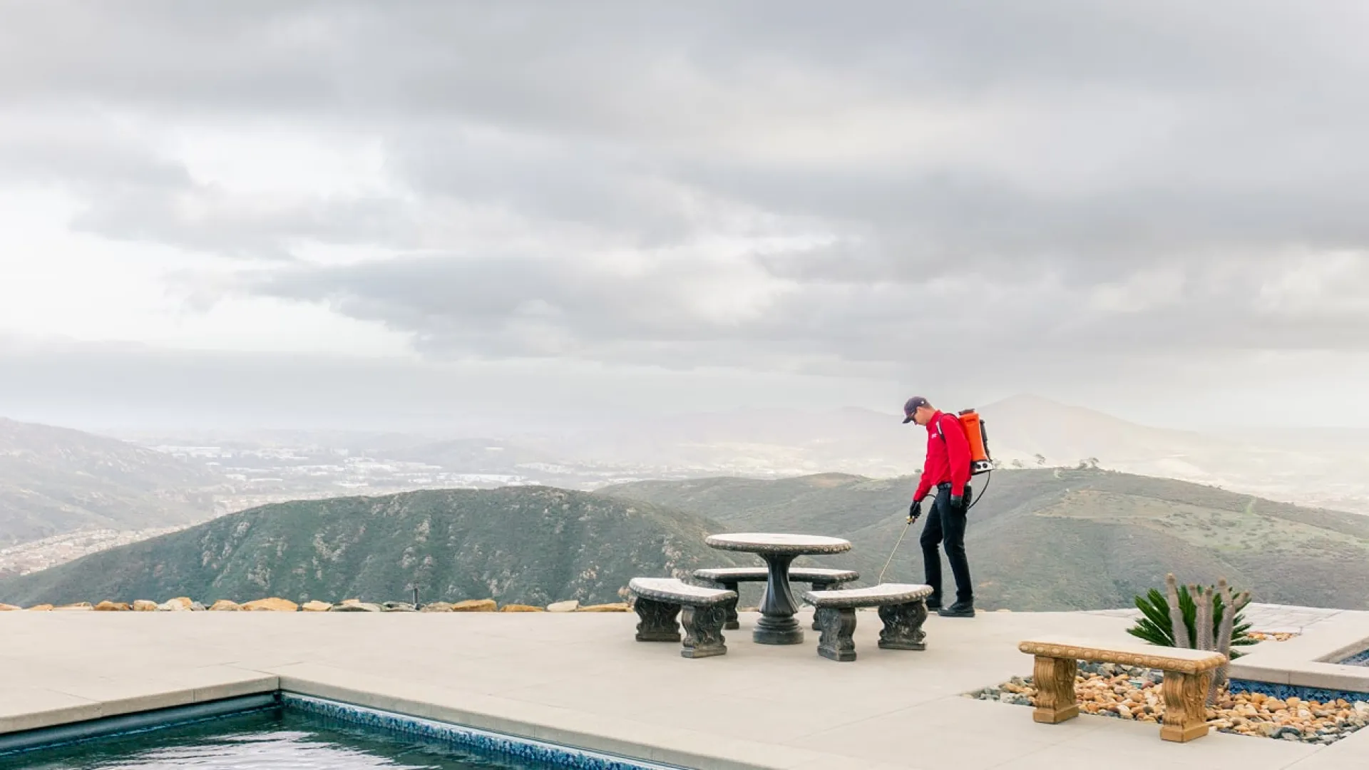 Person in red jacket near outdoor stone table by pool with mountains under cloudy sky in background