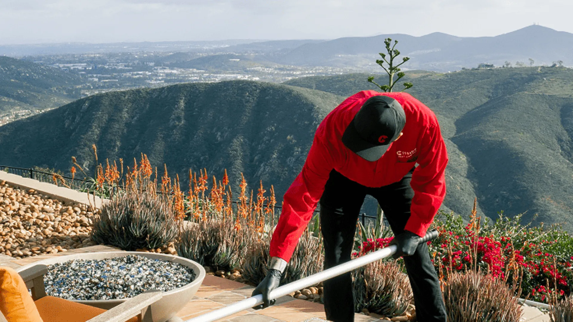 Gardener in red jacket tending plants on patio with mountain views and desert landscaping under clear sky
