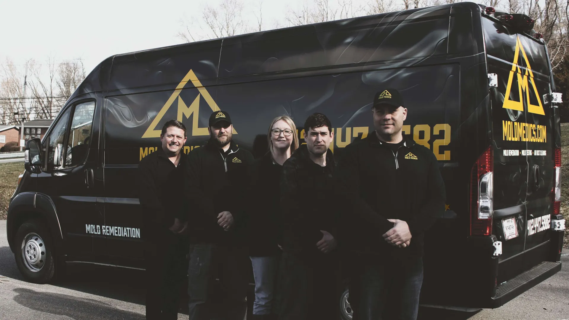 Five Mold Medics team members standing in front of a black mold remediation van with company logo.