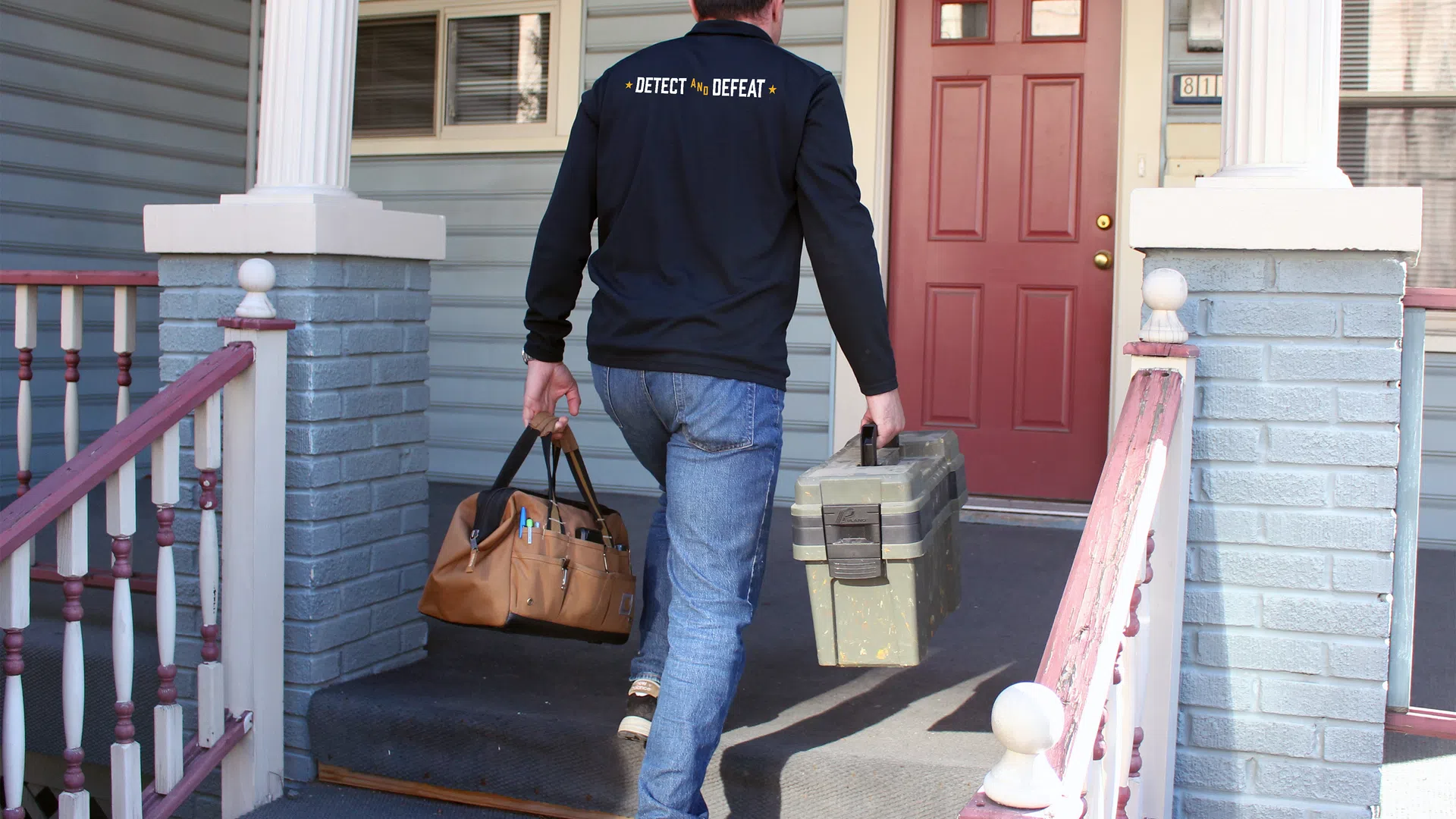 Man wearing Detect and Defeat jacket carrying tool bags up porch steps to a red front door of a house.