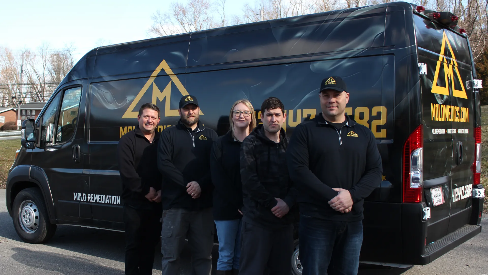 Group of five mold remediation workers standing in front of a black company van with yellow logo and contact info.