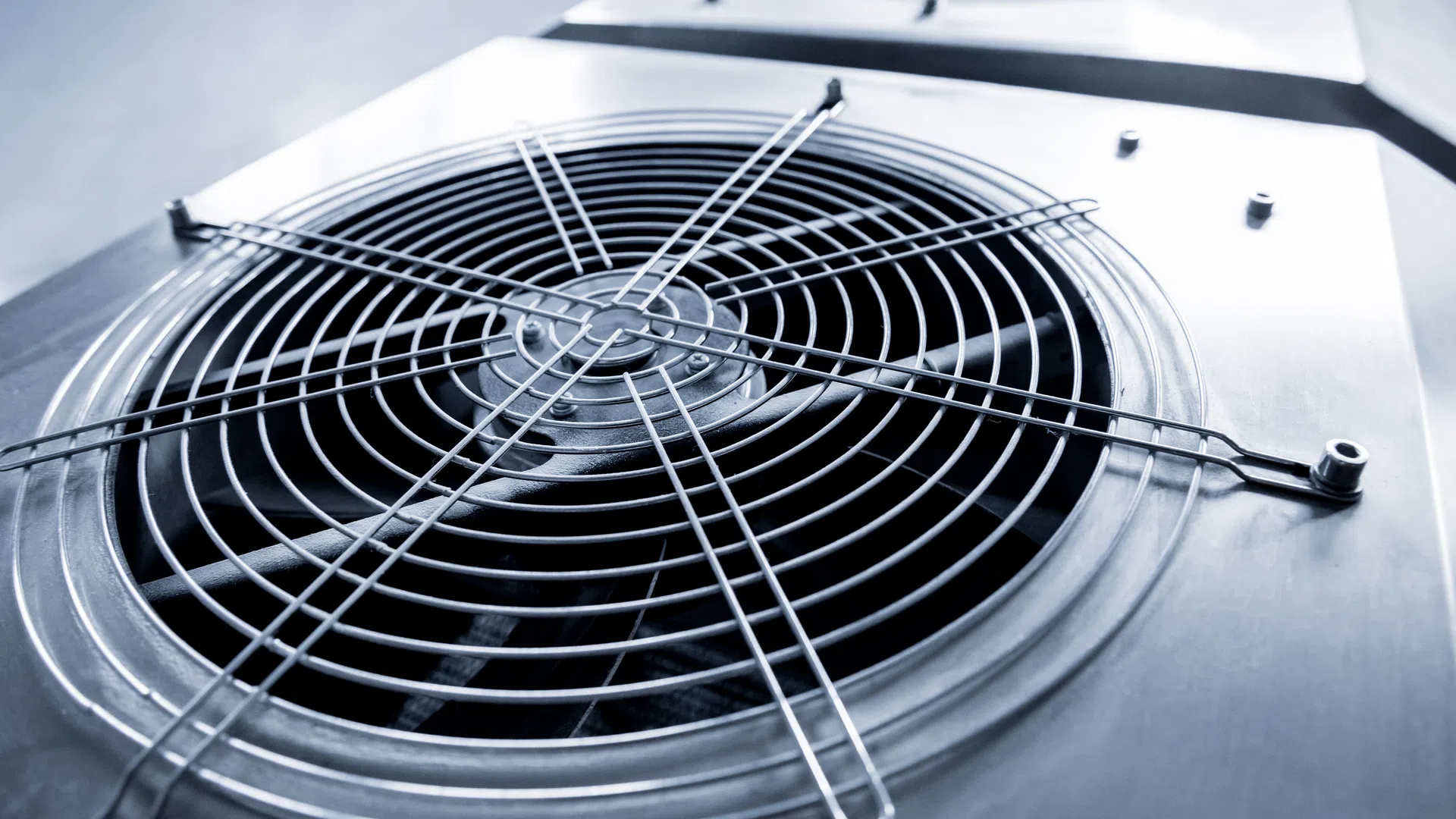 Close-up of industrial air conditioning unit fan with metal grille and blades in a cool blue tone.