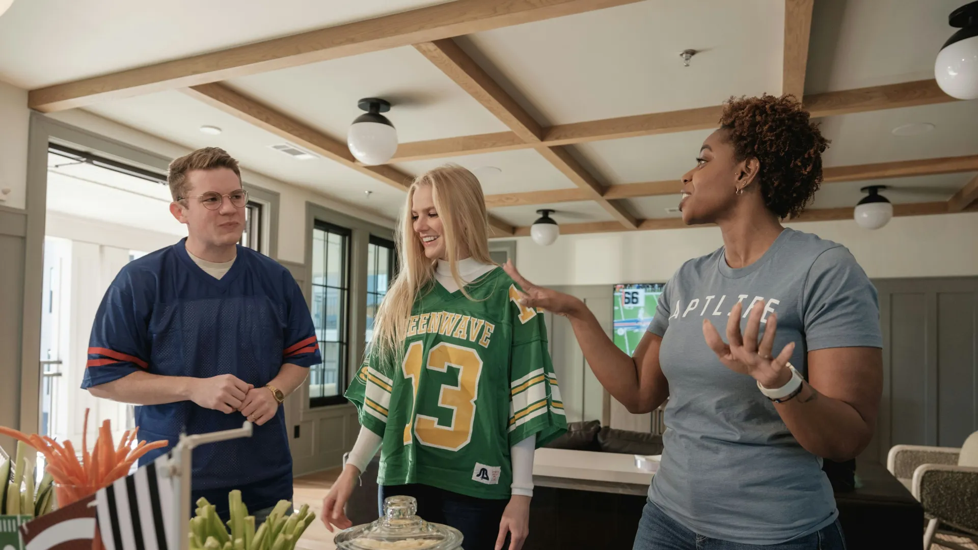 Three friends wearing sports jerseys enjoying a game day gathering with snacks in a cozy living room.