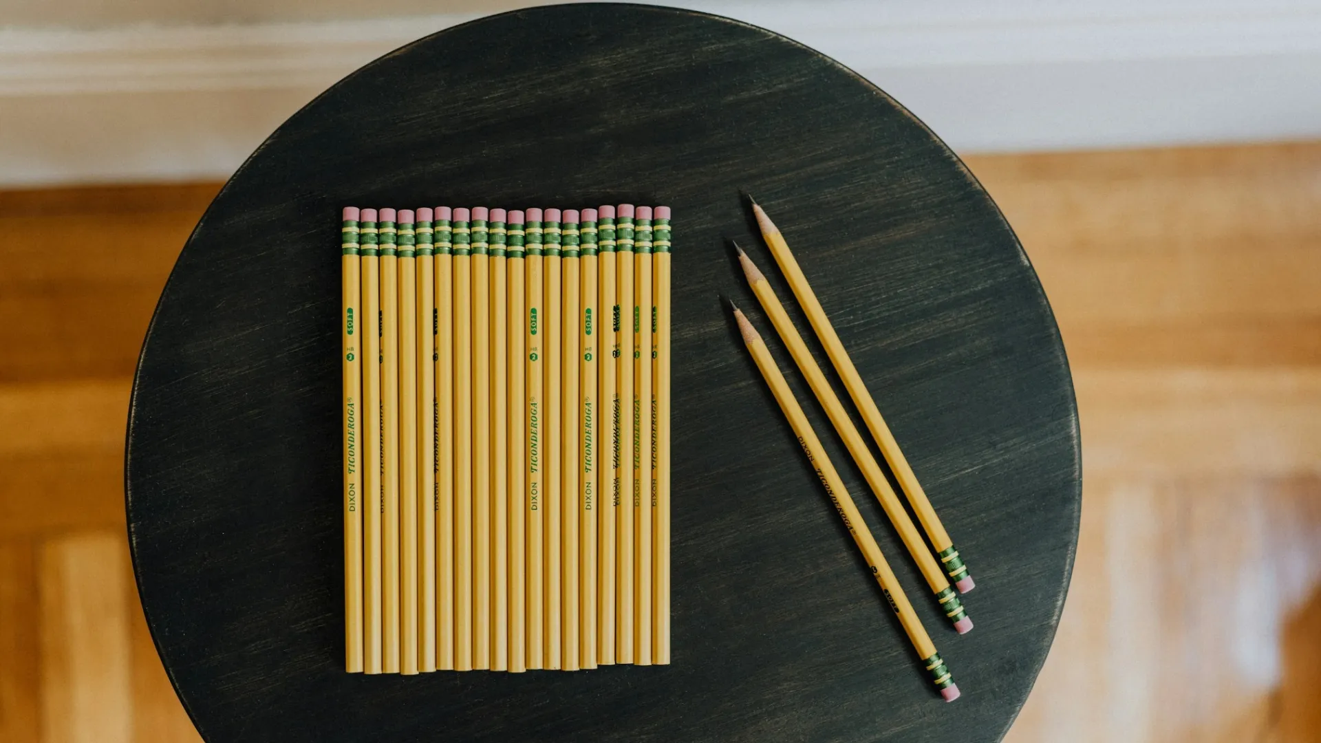 Group of aligned yellow pencils with erasers on round dark wooden table and three scattered pencils