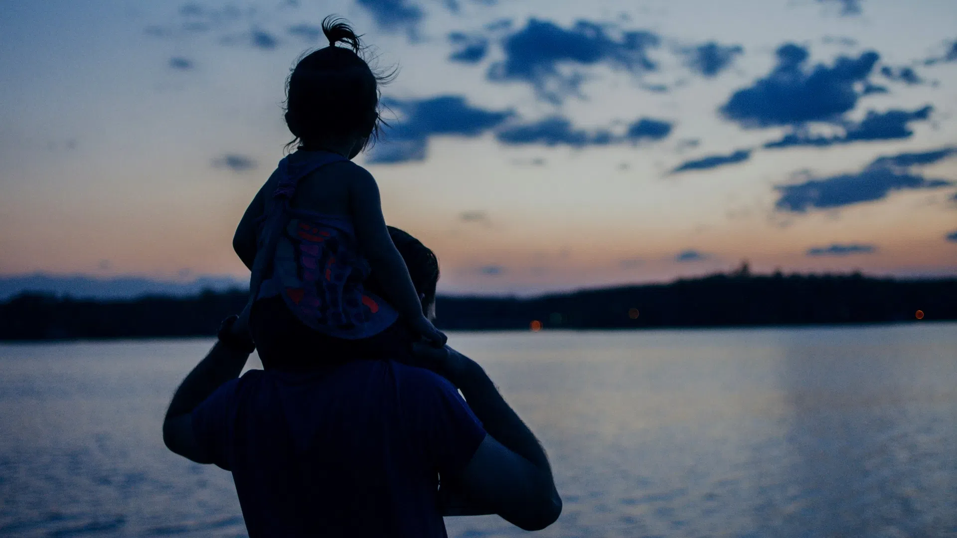 Child sitting on father's shoulders by a lake during a peaceful sunset with calm water and evening sky.