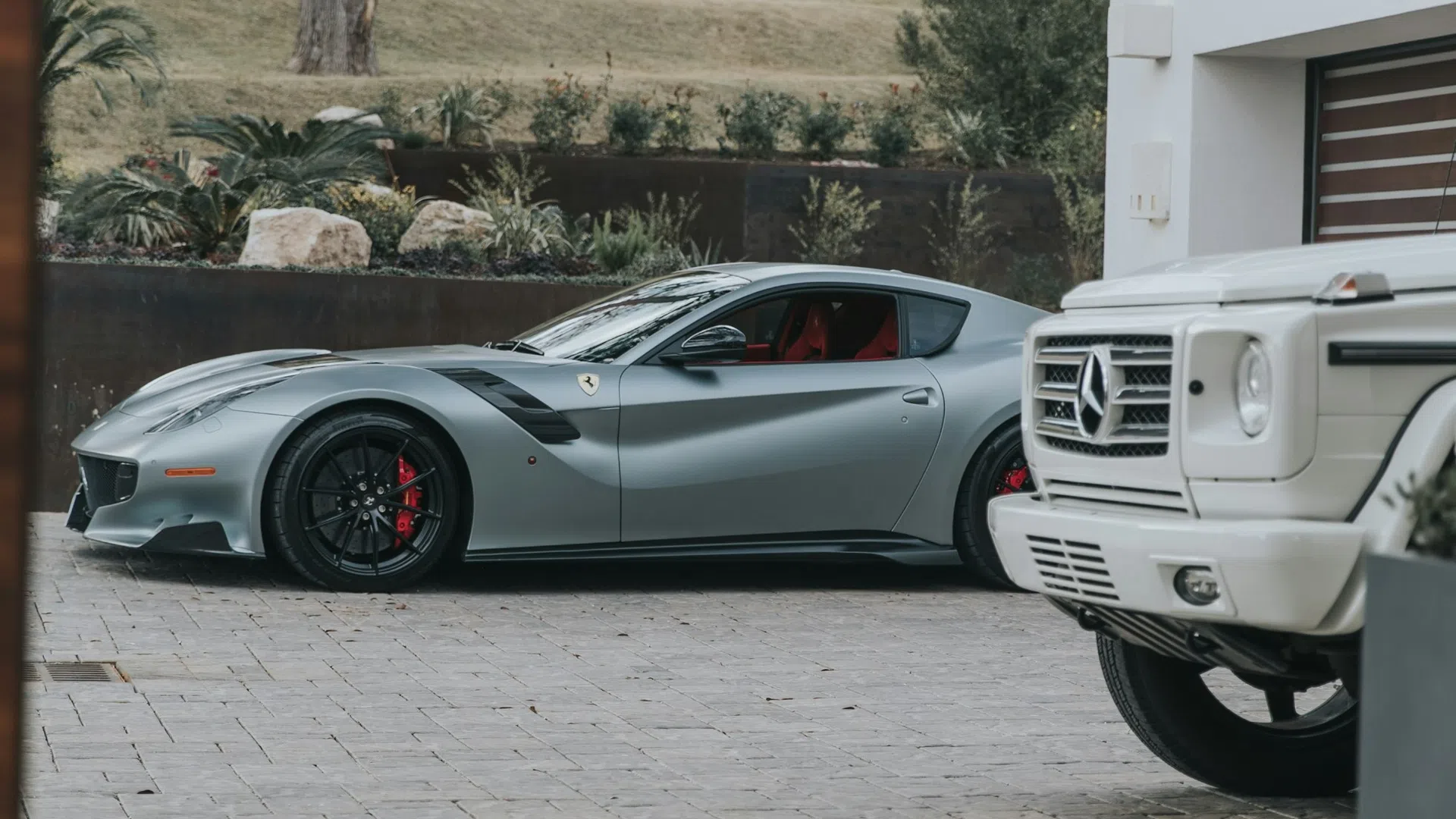 Silver Ferrari sports car and white Mercedes SUV parked in a luxury driveway with greenery in the background
