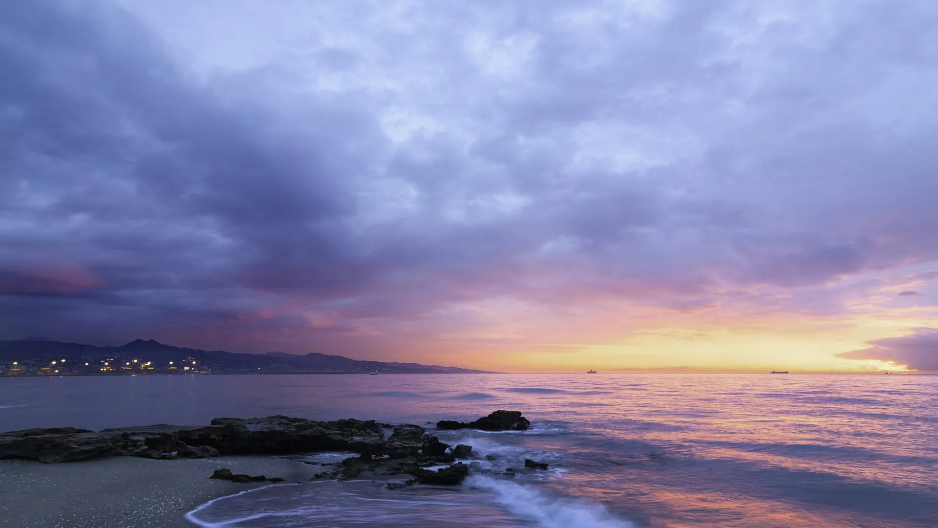 Sunset over a rocky beach with calm waves, colorful sky, and distant mountains under cloudy weather.