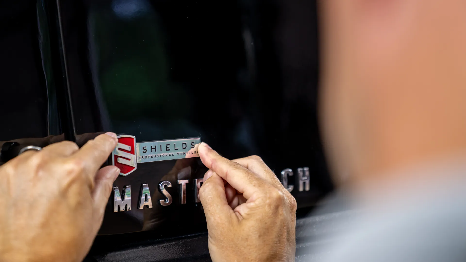 Person applying a Shields professional vehicles logo sticker on a black vehicle surface near MASTER TECH lettering.
