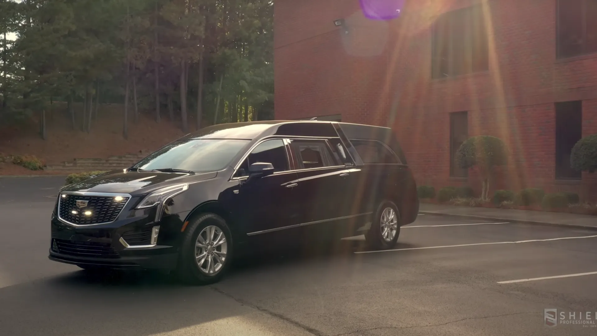 Black Cadillac XT5 hearse parked in front of a brick building with sunlight flaring.