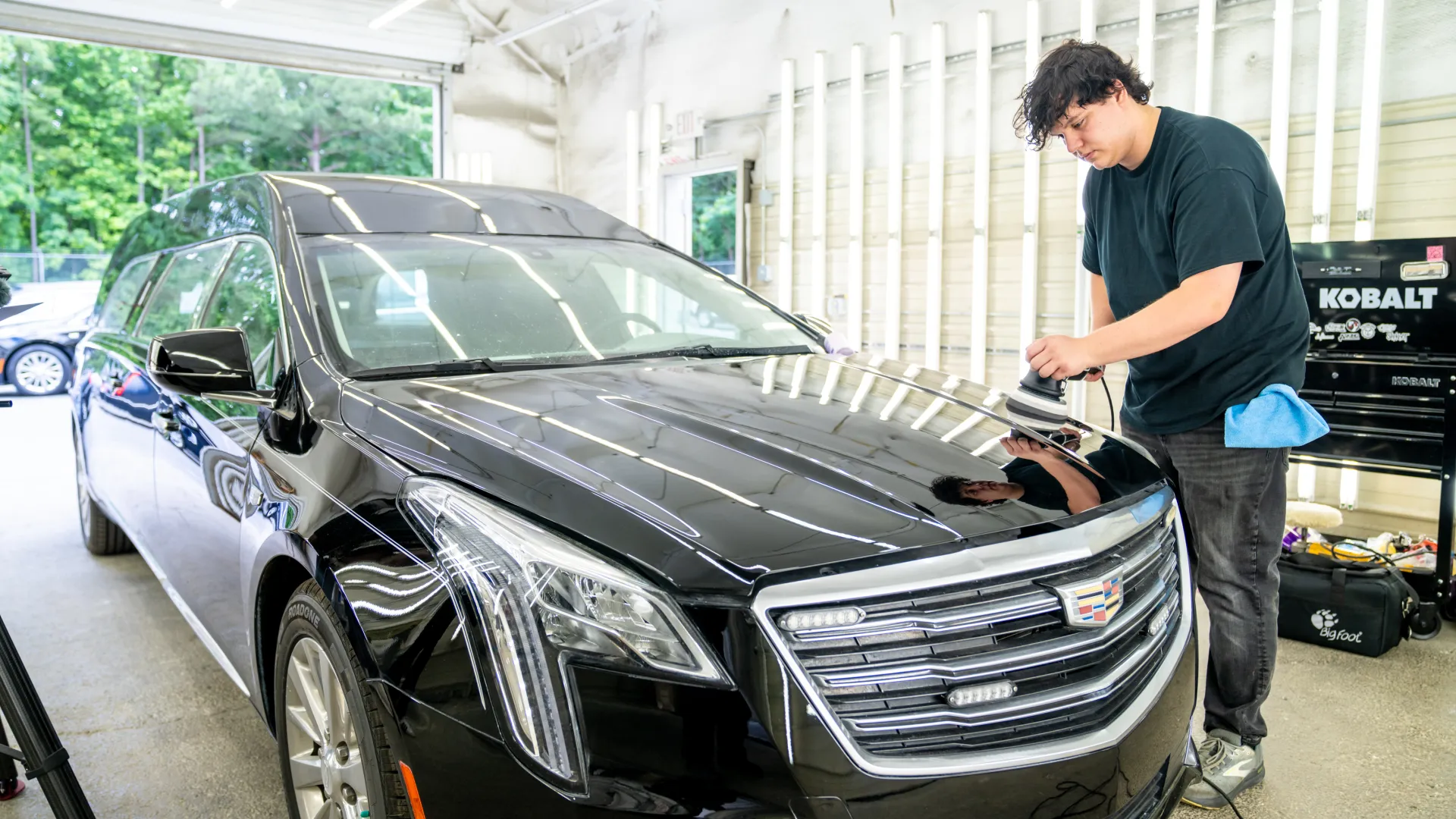 Man polishing the hood of a black Cadillac limousine inside a bright garage with large windows.