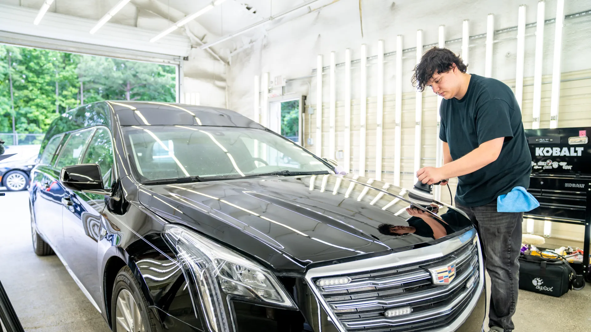Man polishing the hood of a black Cadillac limousine inside a bright garage with large windows.