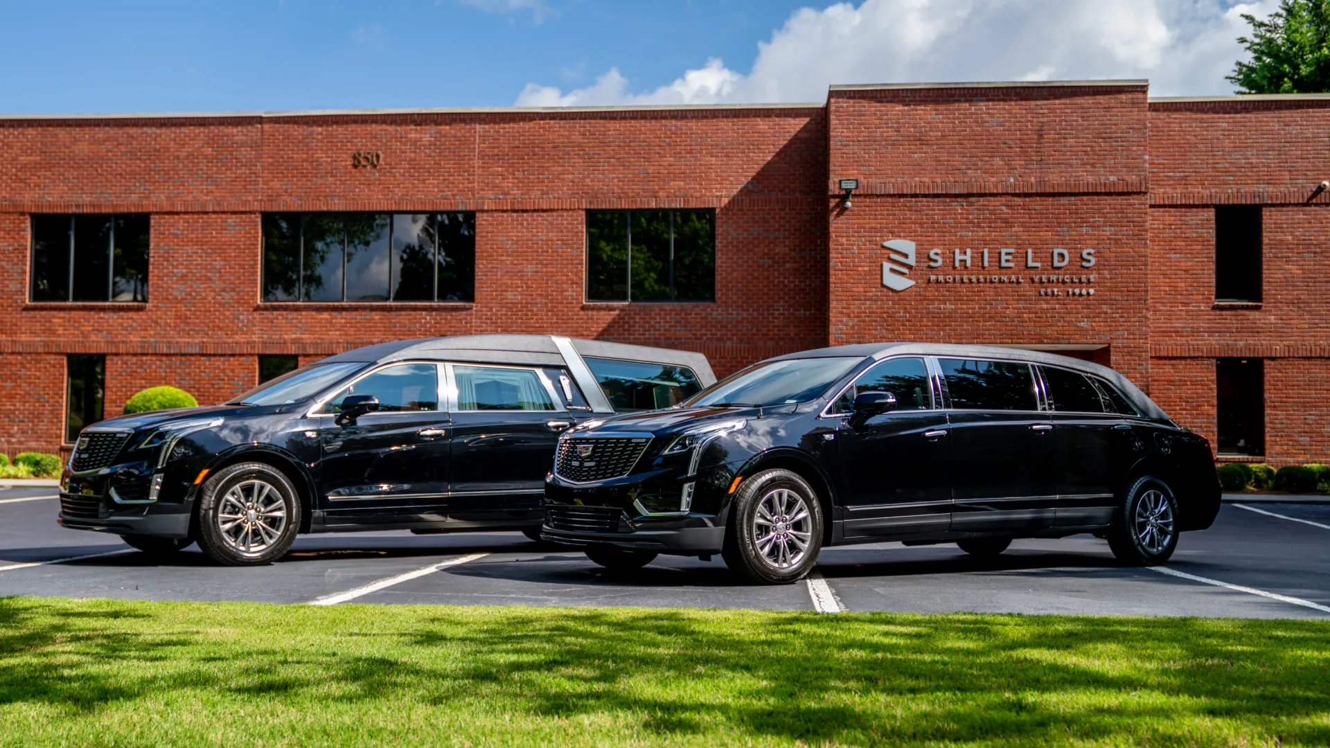 Two sleek black hearses parked in front of a red brick building with green grass and blue sky.