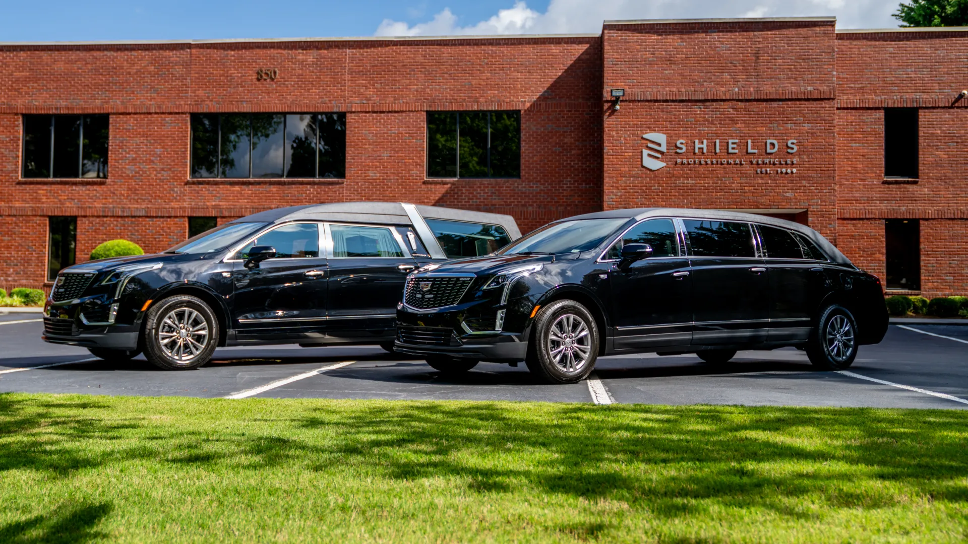 Two sleek black hearses parked in front of a red brick building with green grass and blue sky.