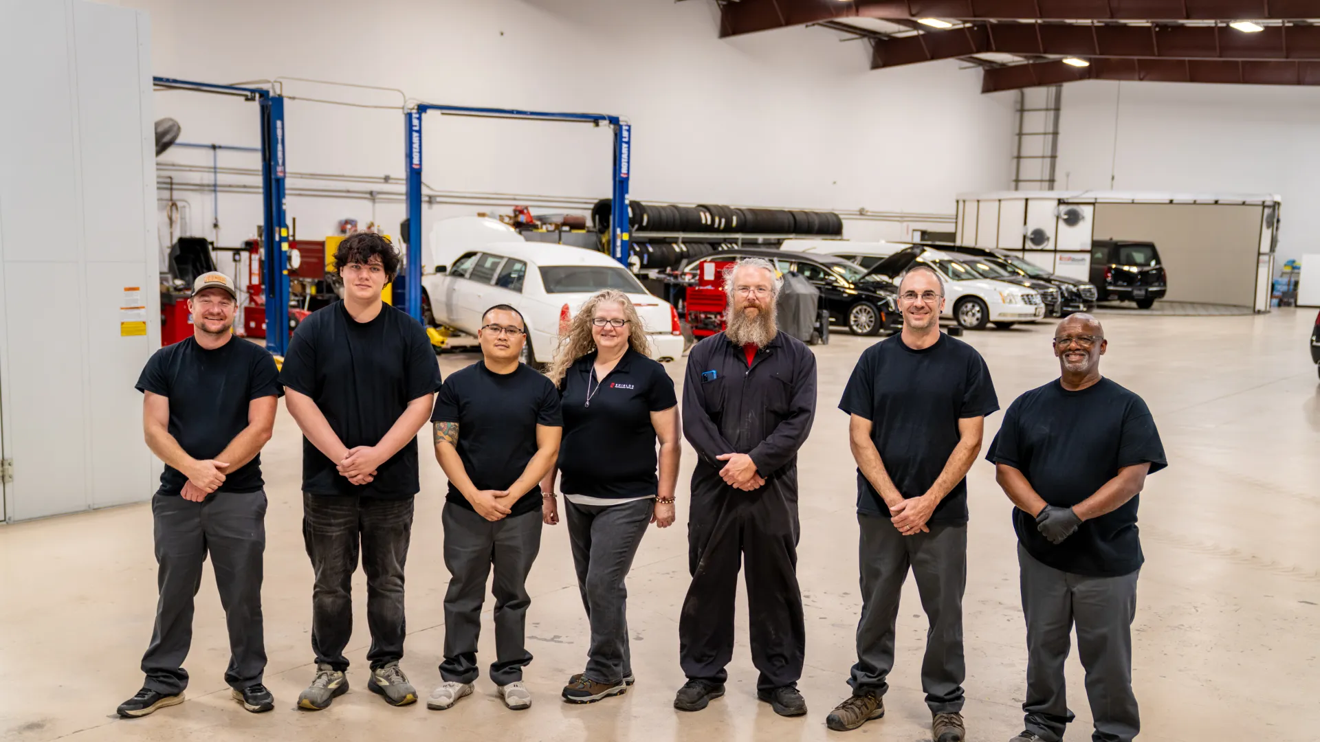 Seven auto repair technicians stand in a spacious garage with cars and equipment in the background.