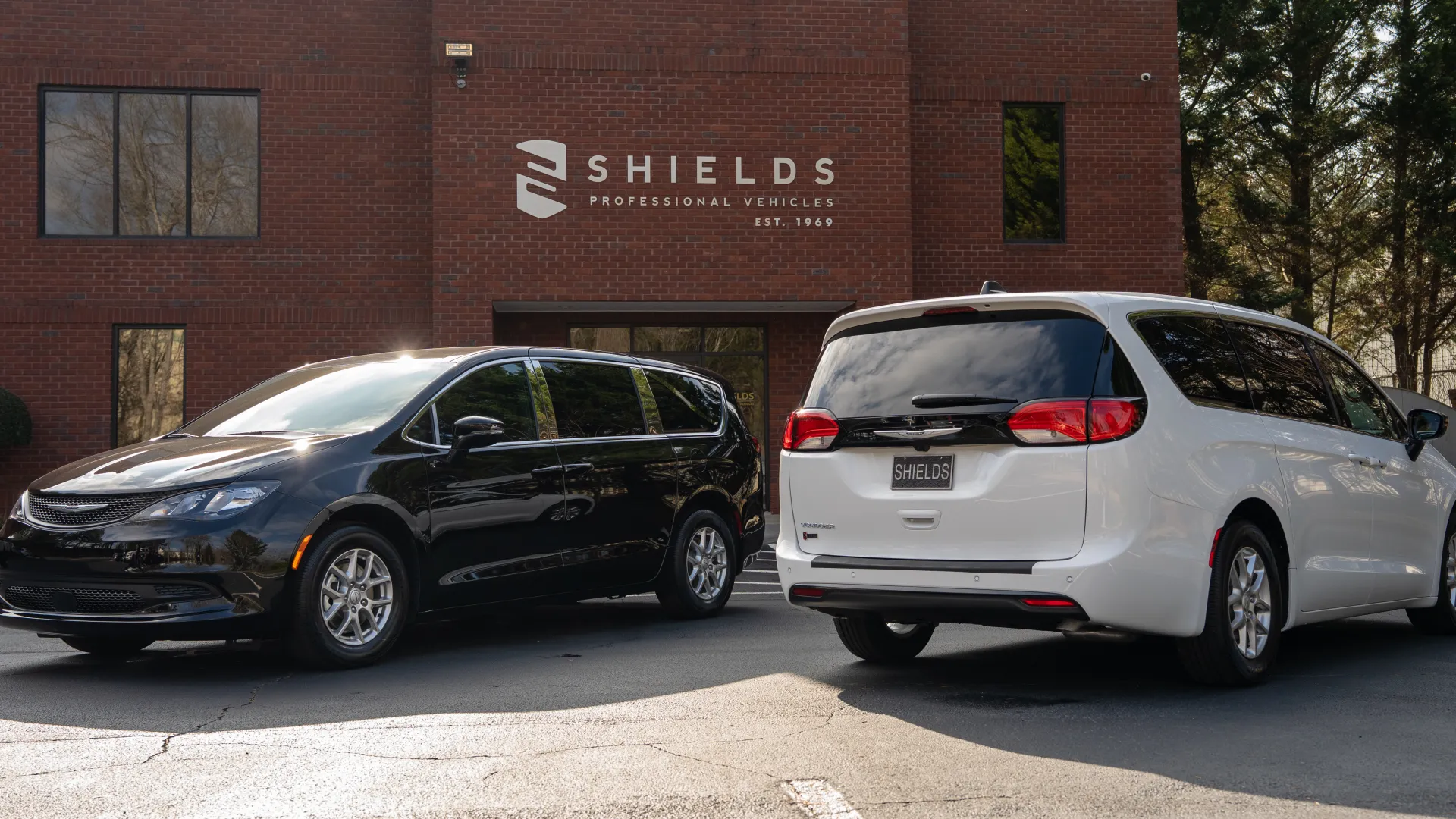 Black and white minivans parked outside Shields Professional Vehicles building with clear logo on brick wall.
