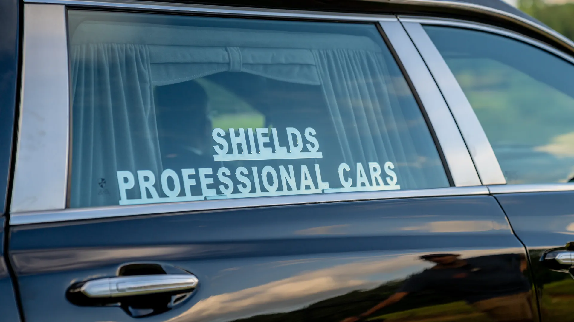Black car window with white lettering reading Shields Professional Cars and white curtains inside.
