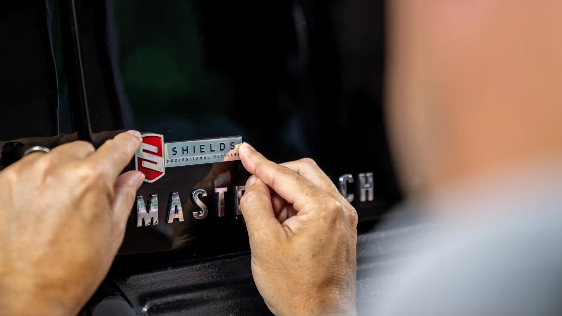 Person applying a Shields Professional Vehicles badge decal to a black vehicle near the MasterTech emblem.