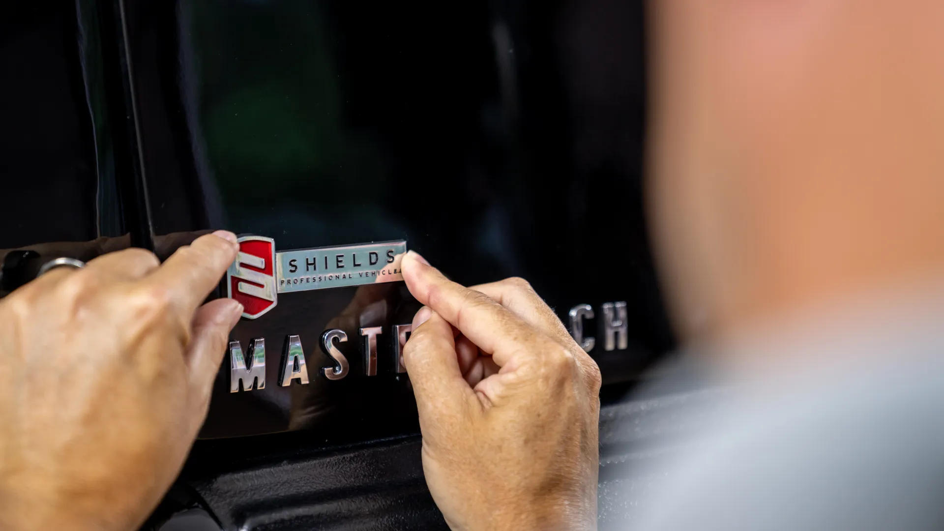 Person applying a Shields Professional Vehicles badge decal to a black vehicle near the MasterTech emblem.