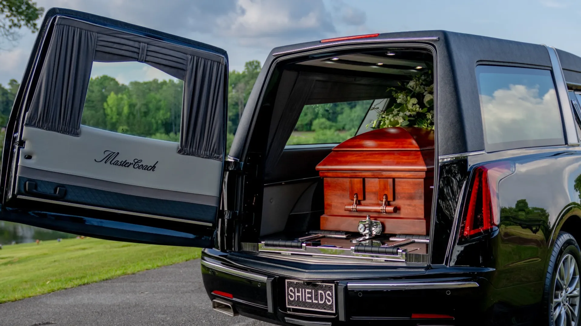 Black hearse with open rear door revealing a wooden casket inside on a road beside green trees and blue sky.