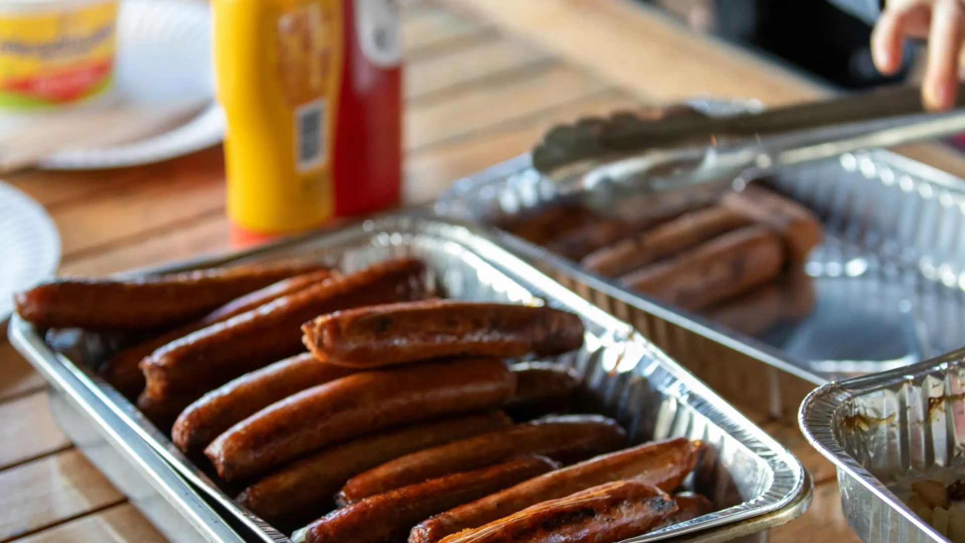 Grilled sausages in aluminum trays with ketchup and mustard bottles on wooden table at outdoor meal.