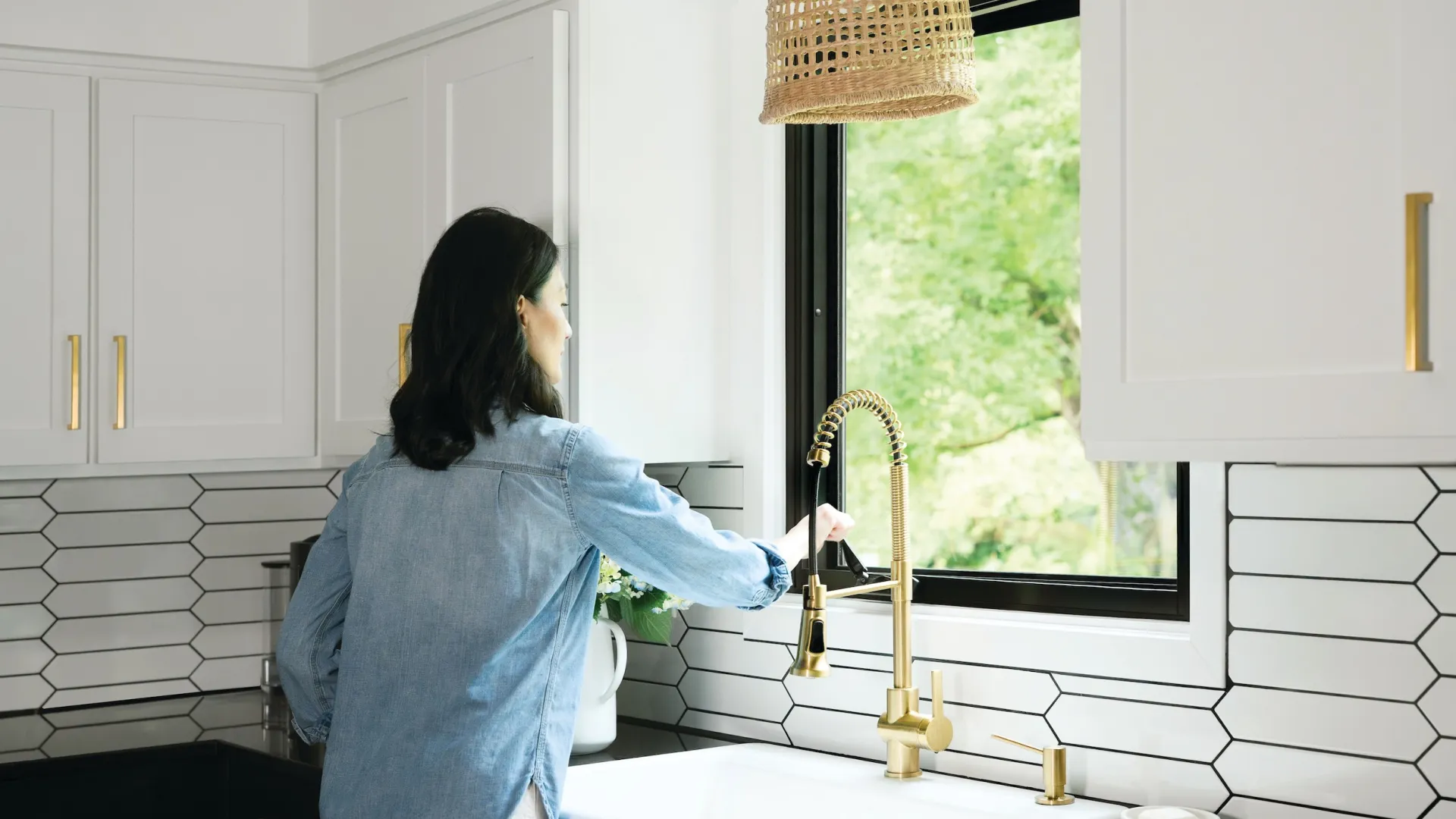 Woman in denim shirt uses modern gold kitchen faucet by window in stylish white and black kitchen