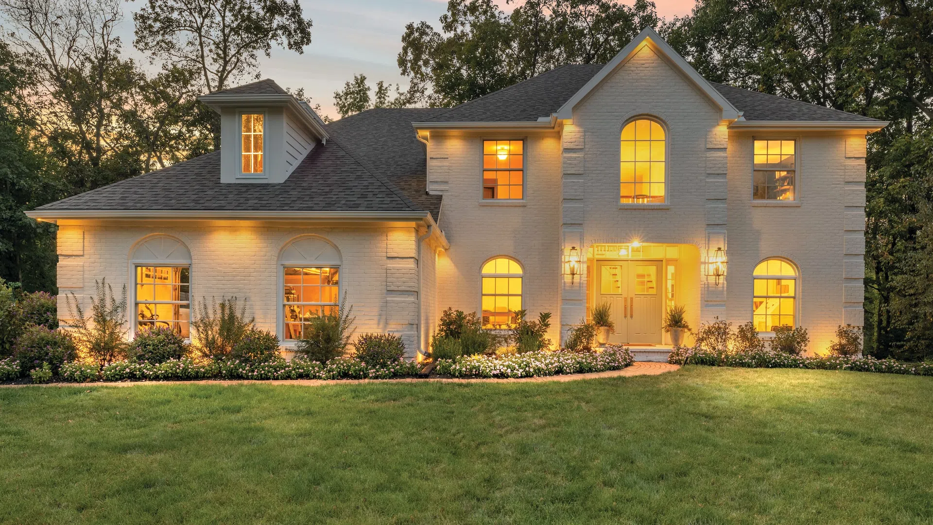 Elegant two-story white brick house with warm interior lights, surrounded by greenery during sunset.
