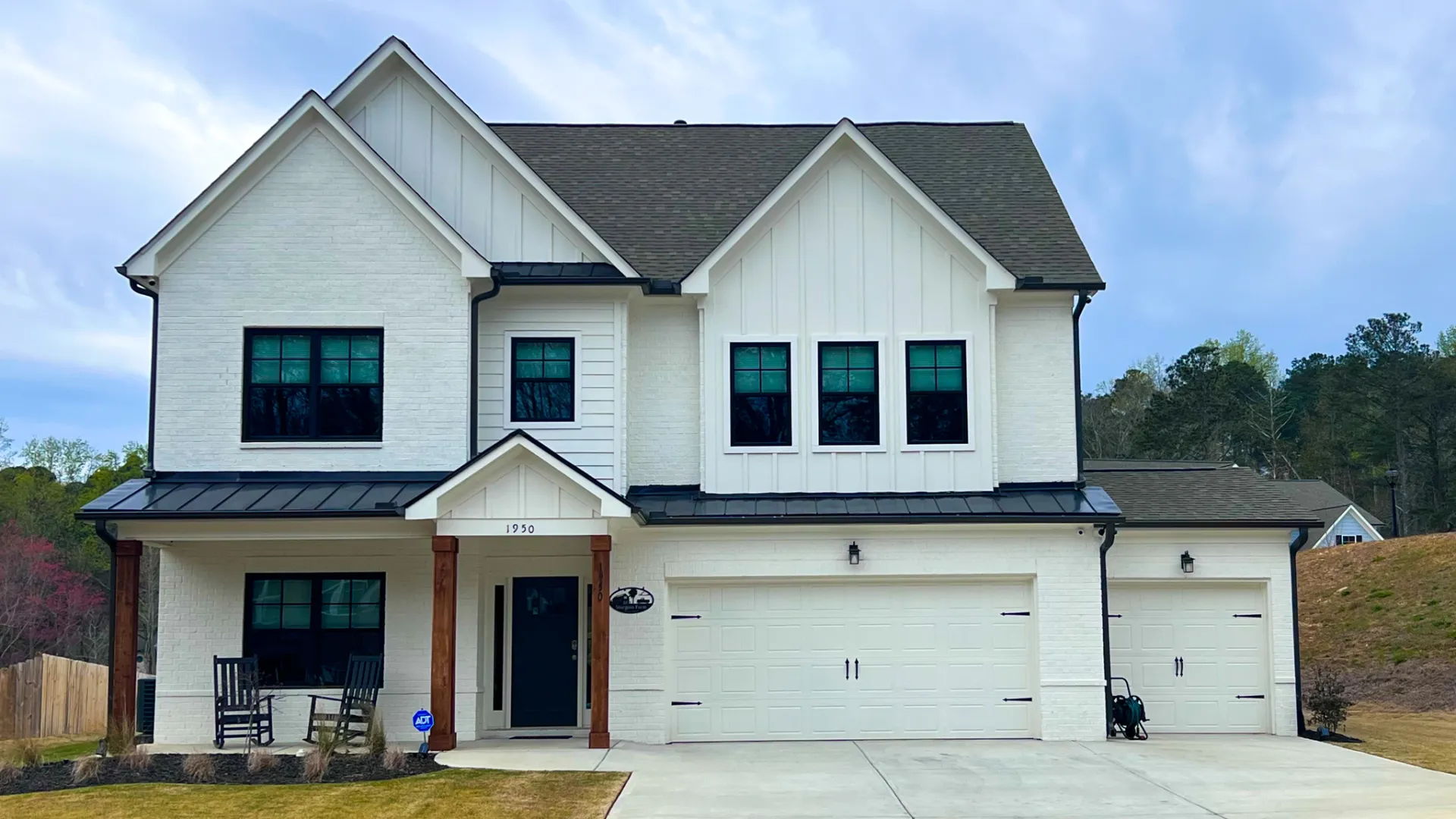 Modern two-story white house with black roof, garage doors, and front porch with rocking chairs under cloudy sky.