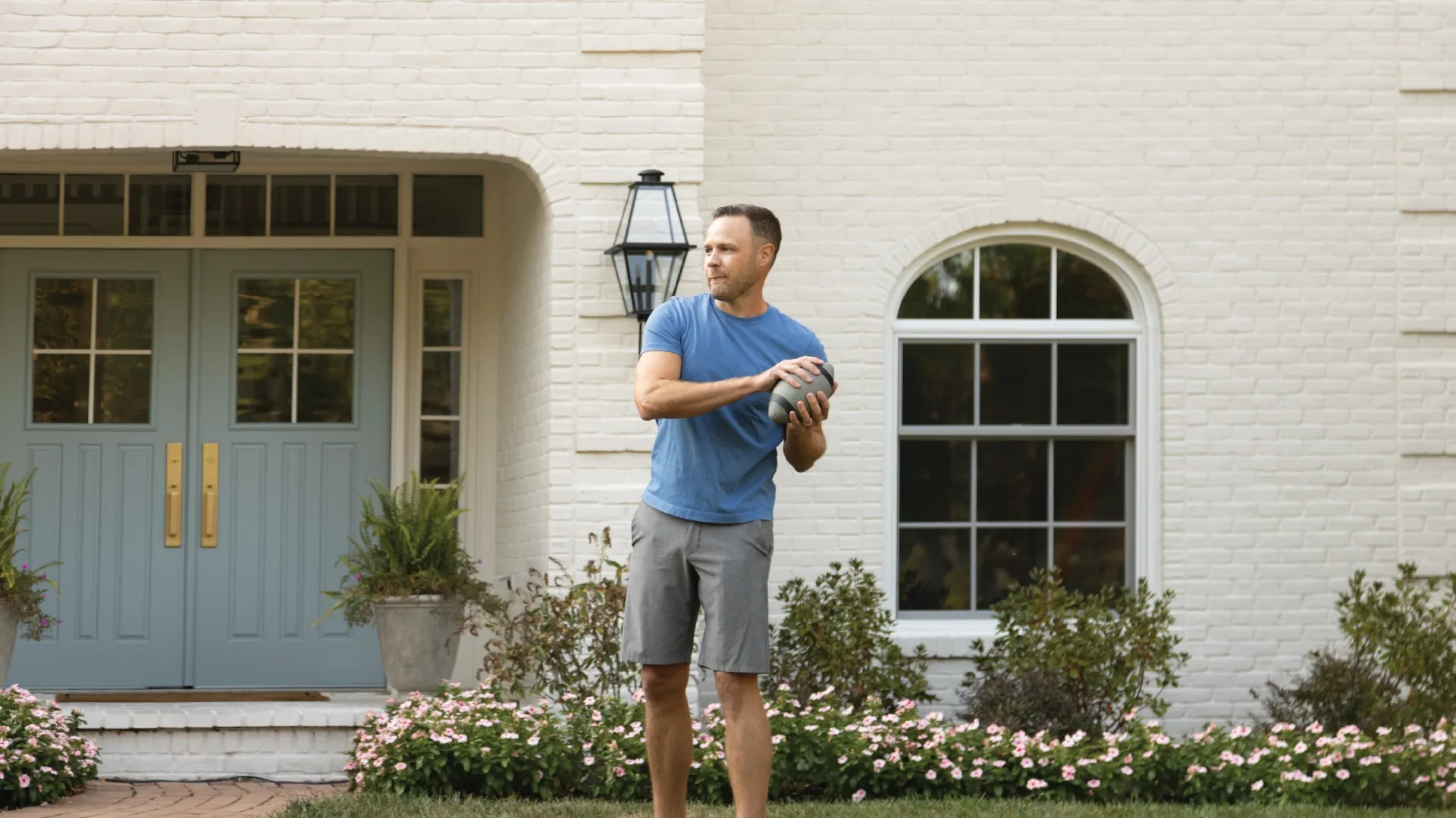 Man in blue t-shirt and gray shorts preparing to throw football outside house with blue doors and flowers.