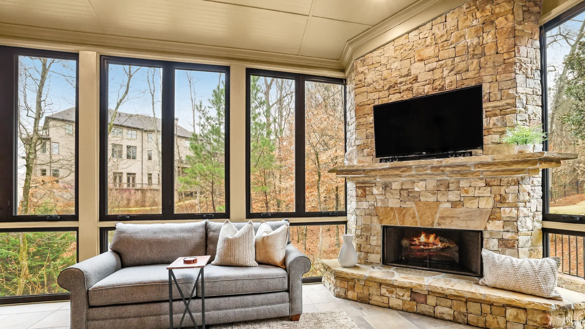 Cozy sunroom with gray sofa, stone fireplace, large windows, and natural view outside