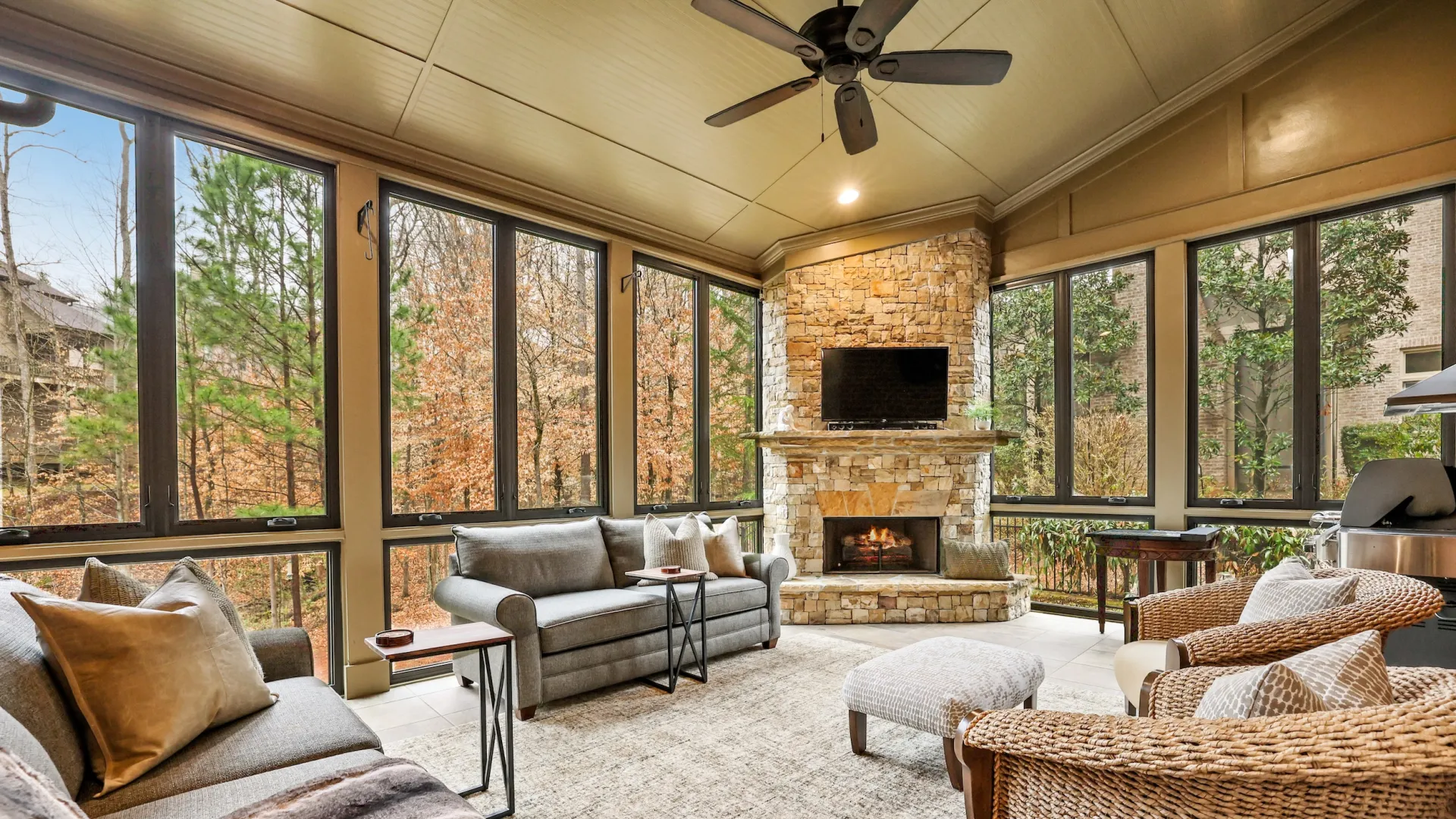 An enclosed porch with stone fireplace, ceiling fan, and seating area with bronze casement windows filling the wall space for a beautiful view of the trees outside.