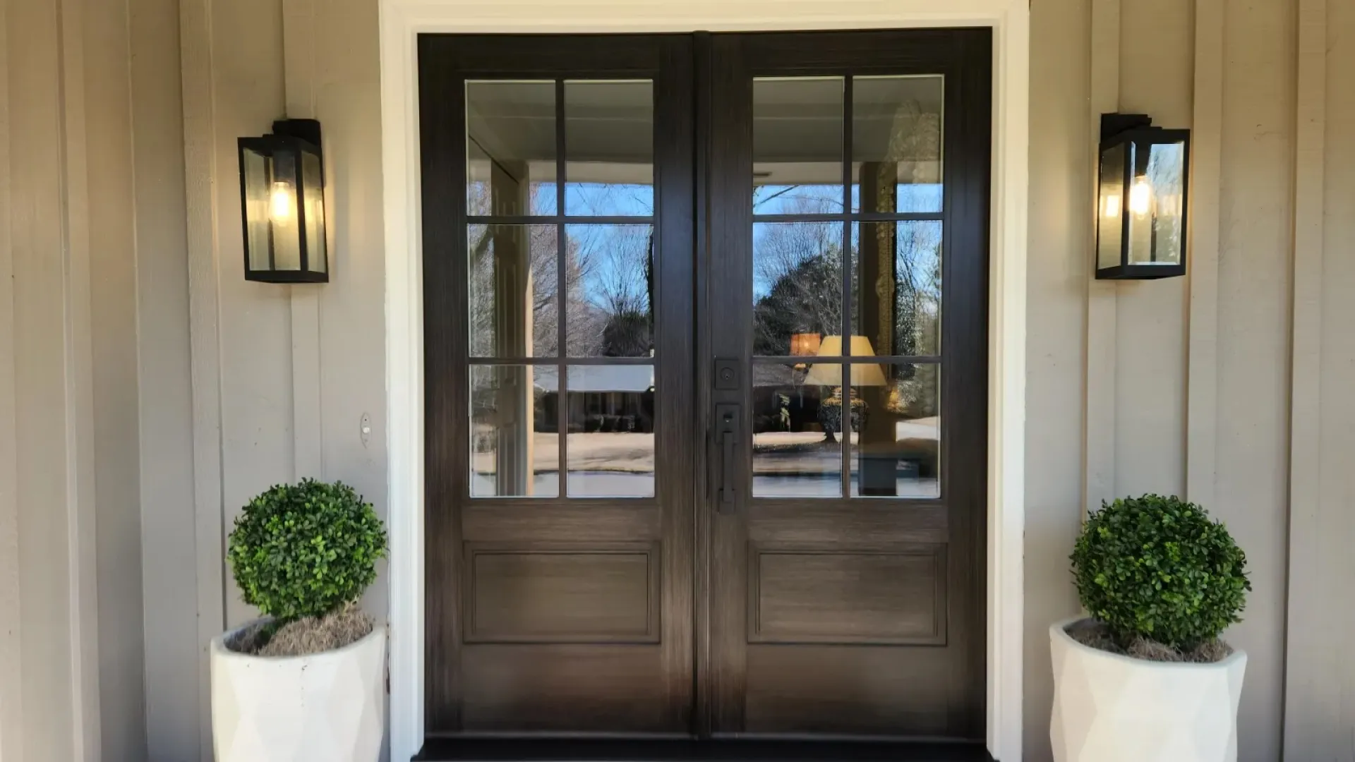 Modern double wooden front doors with glass panels, flanked by wall lanterns and potted plants on porch.