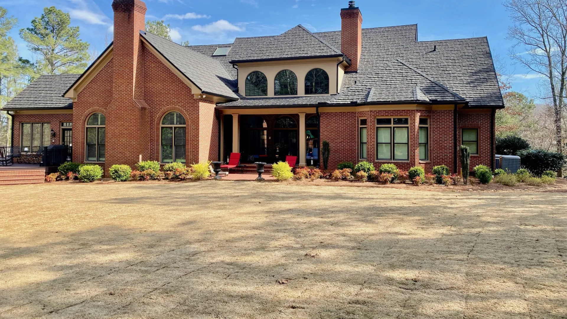 Spacious red brick house with multiple chimneys, arched windows, and a large front yard under a blue sky.