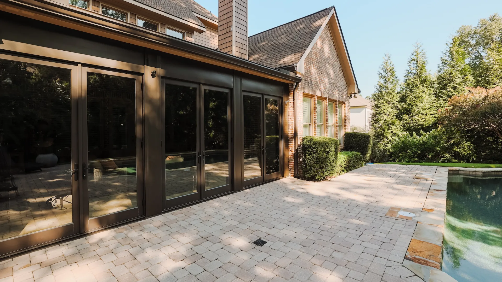 Modern house patio with large glass doors, stone pavers, and a swimming pool surrounded by greenery on a sunny day
