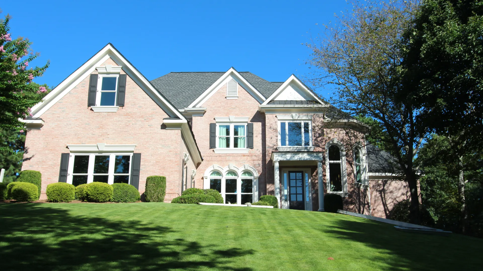 Two-story brick house with large windows, manicured lawn, and surrounding trees under a clear blue sky.