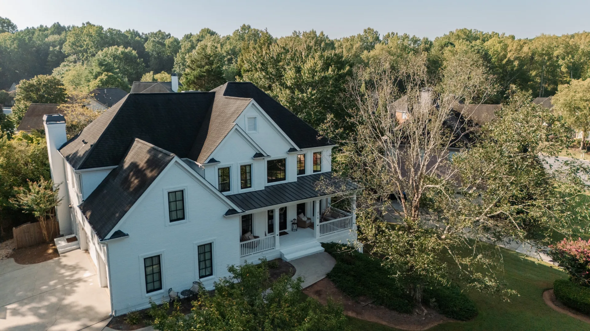 Large white two-story house with black roof surrounded by trees and greenery on a sunny day