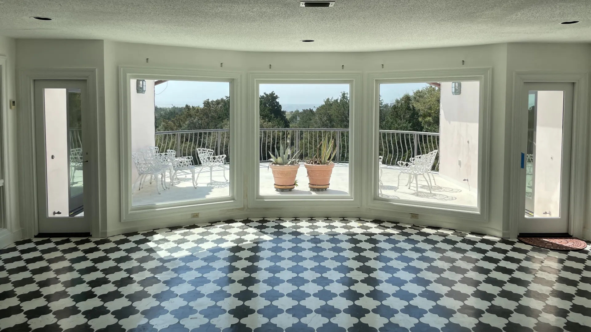 Spacious sunlit room with black and white patterned floor and large windows overlooking outdoor patio with chairs and plants