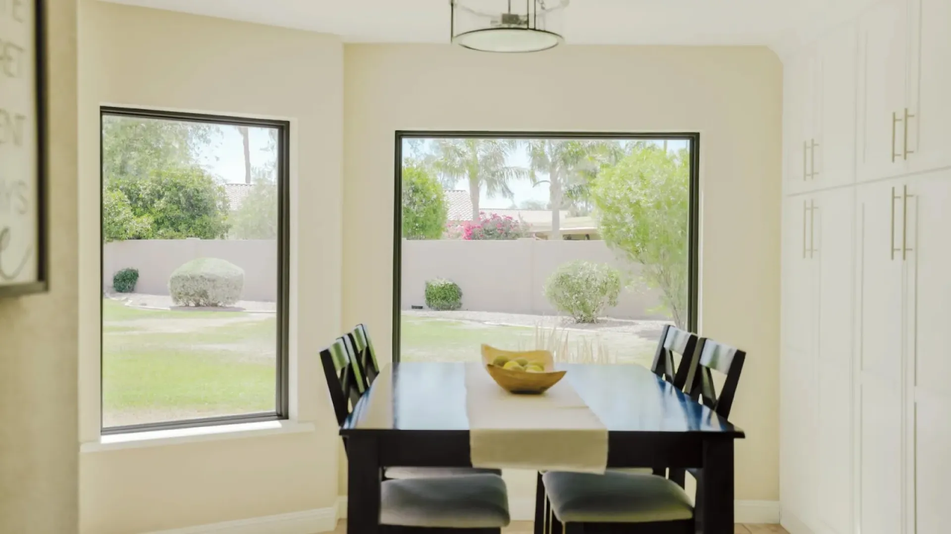 Bright dining area with a dark wooden table, four black chairs, and large windows showing a green garden.