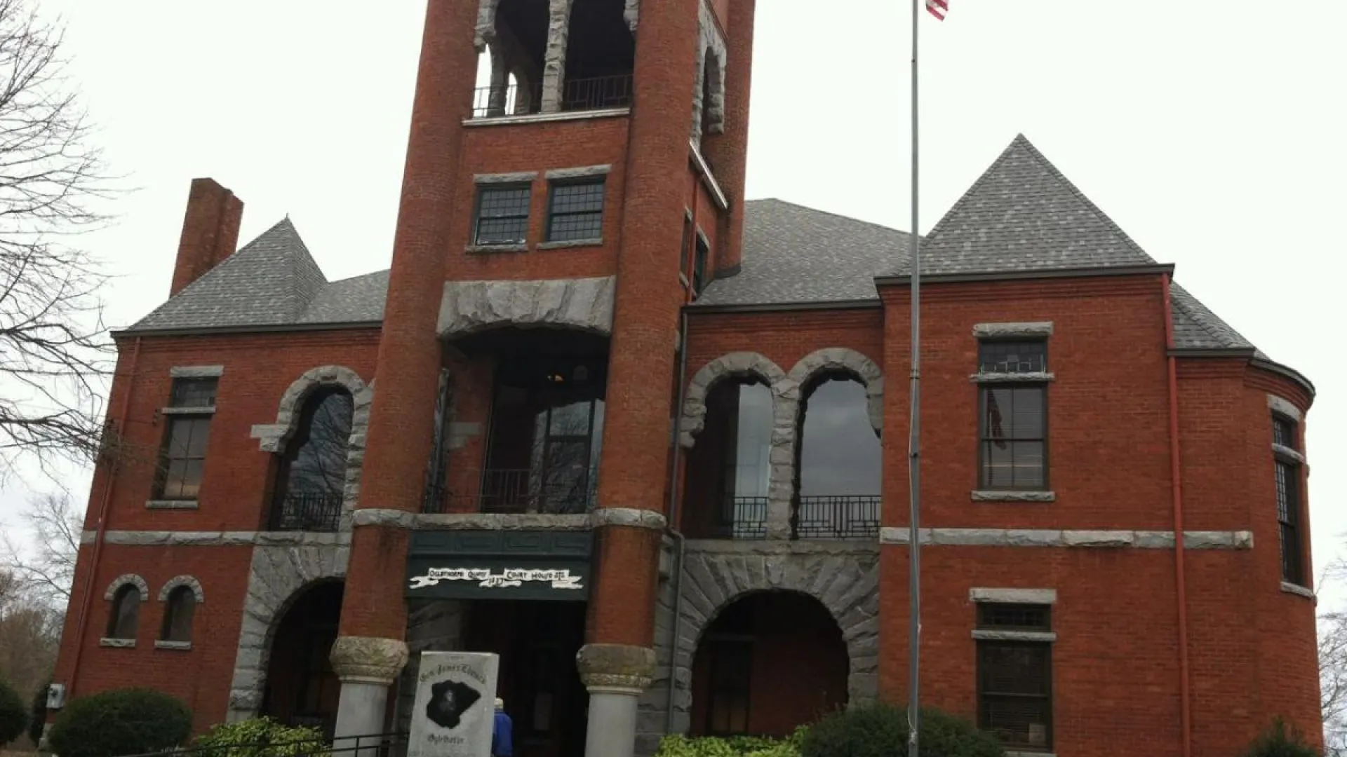 Historic red brick courthouse with clock tower and American flagpole in front on a cloudy day.