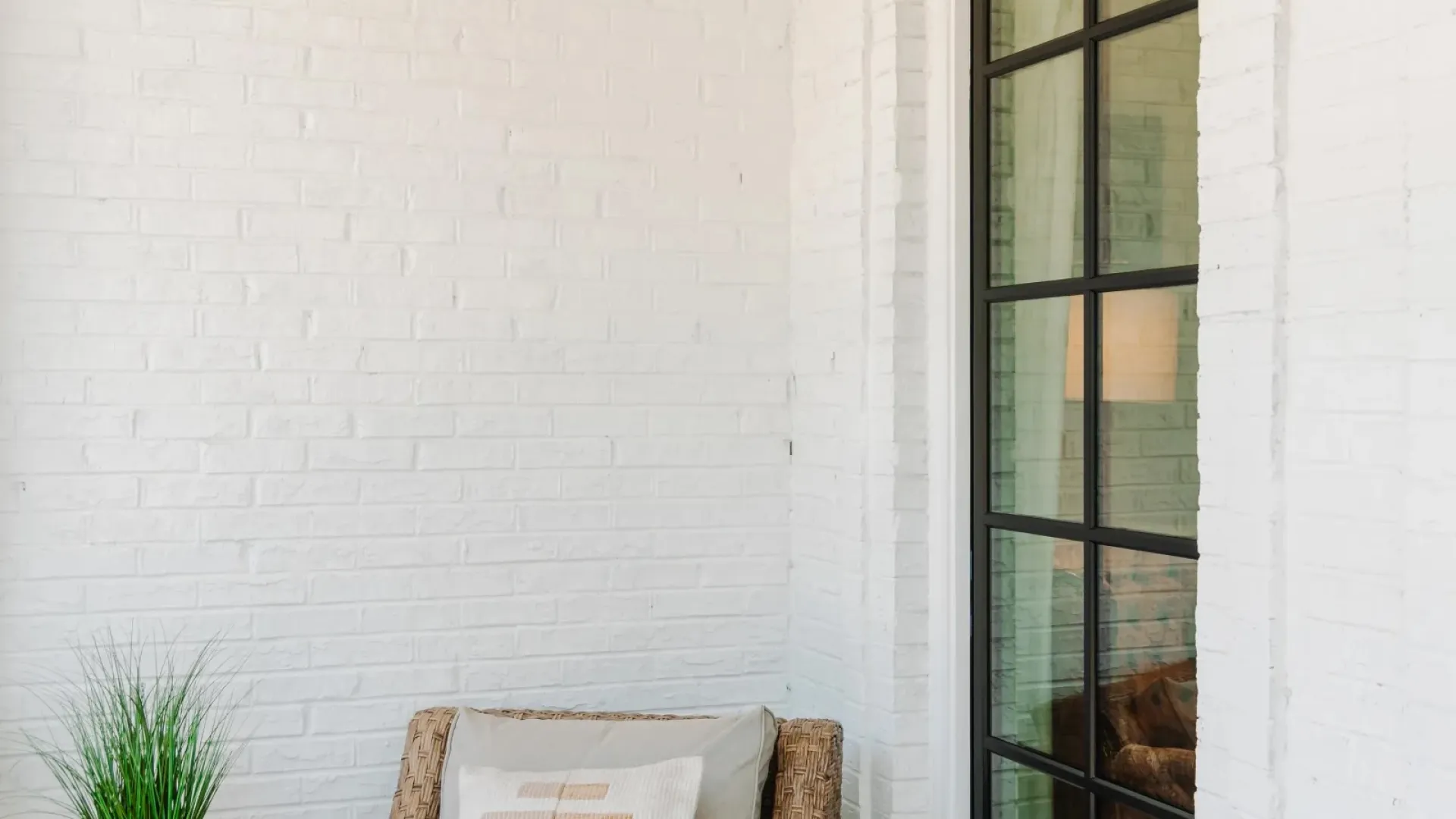Cozy corner with wicker chair, decorative pillow, green plant, and black-framed glass door against white brick wall.