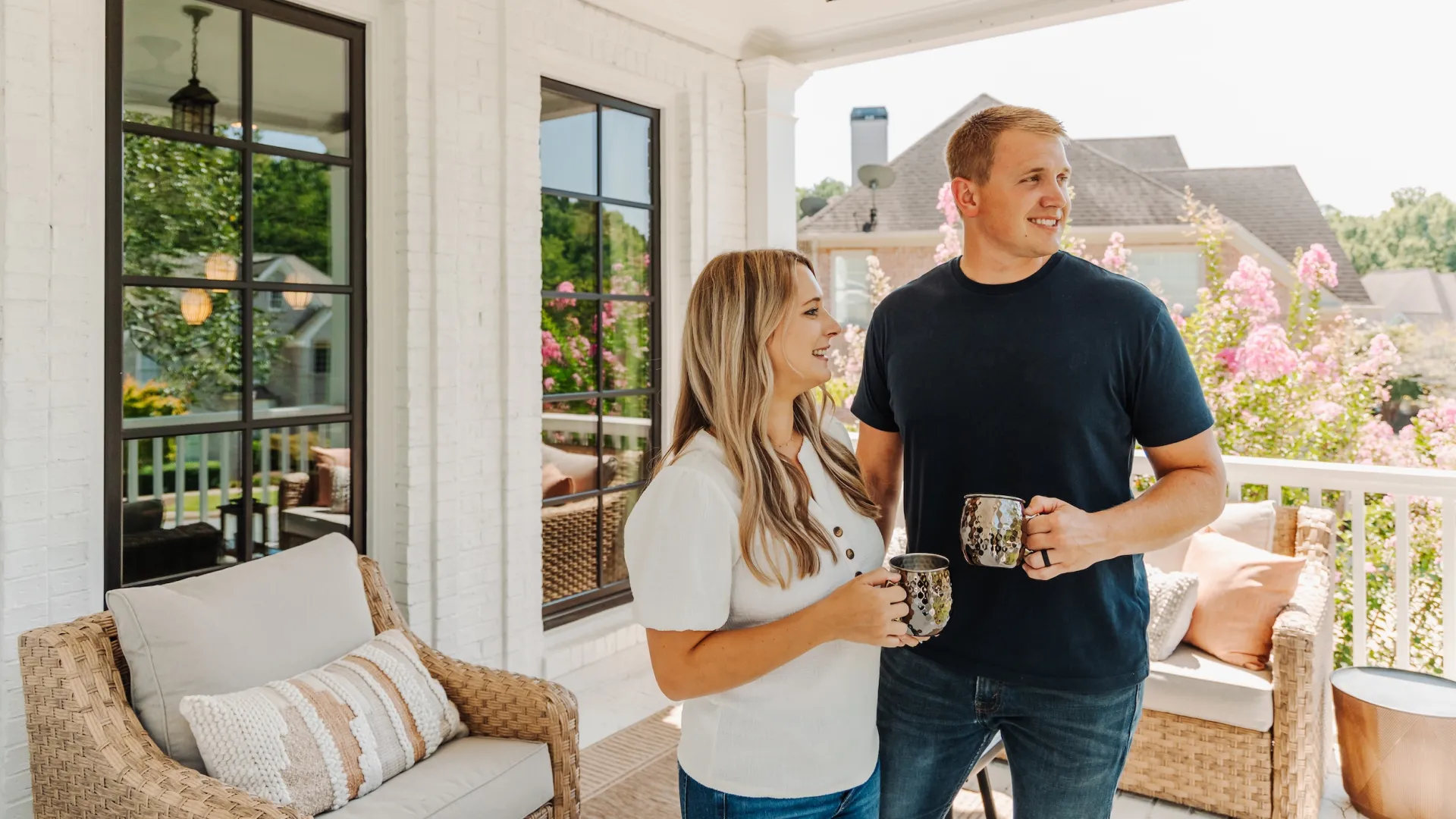 Couple enjoying morning coffee on a cozy porch with wicker furniture and blooming flowers in the background
