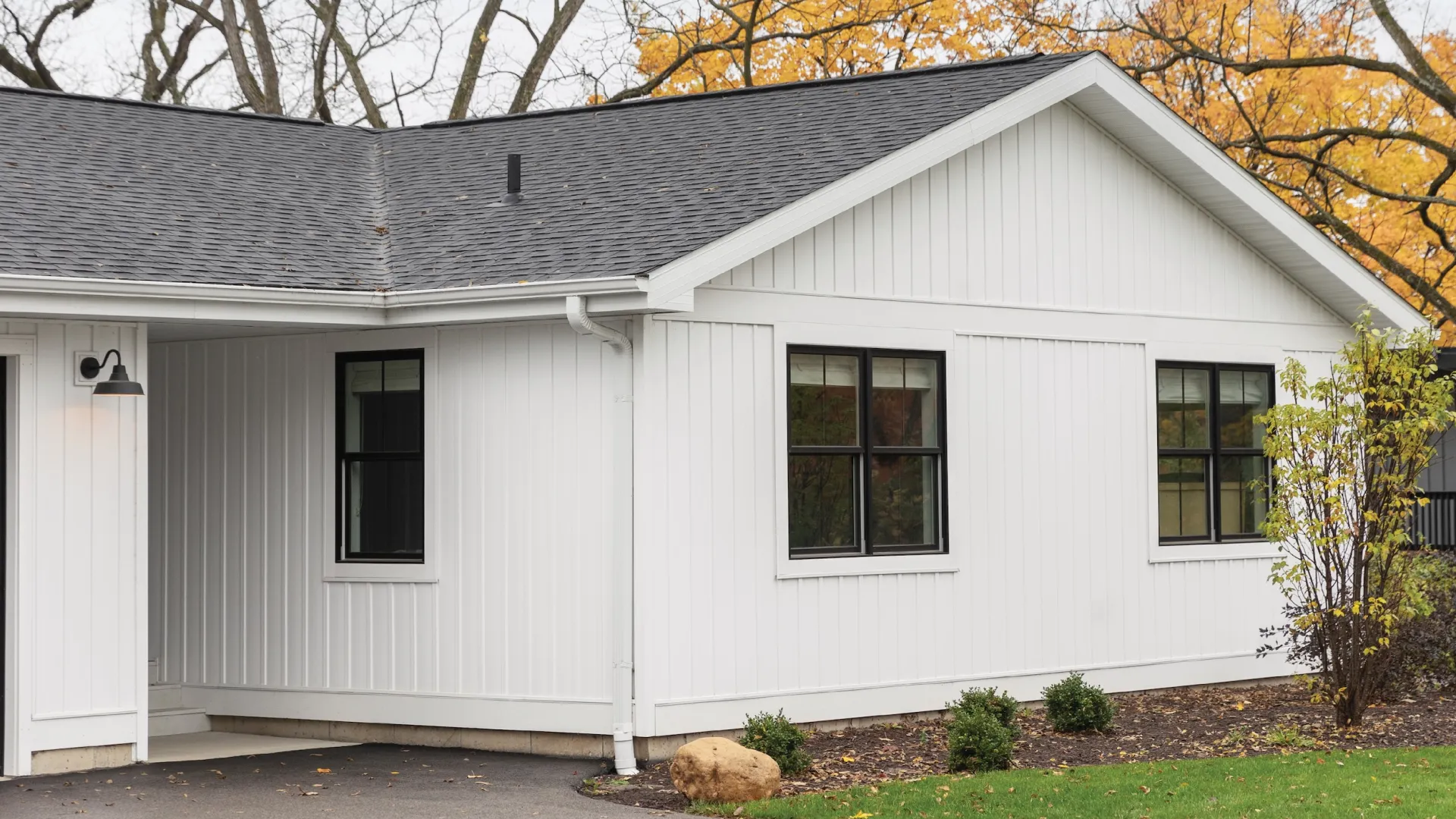 White modern house exterior with black fiberglass double hung window frames, gray roof, fall trees, and minimal landscaping.